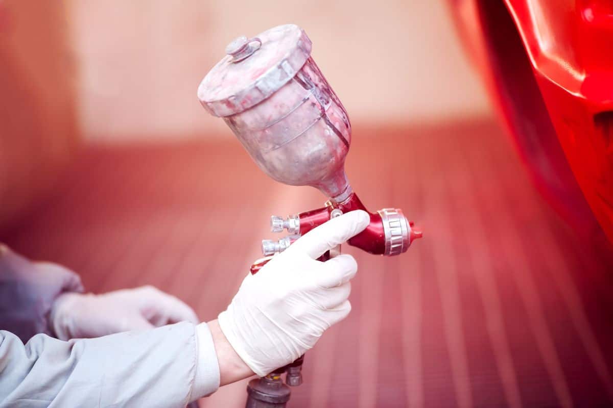 Worker painting a red car in painting booth using professional tools and spray gun.