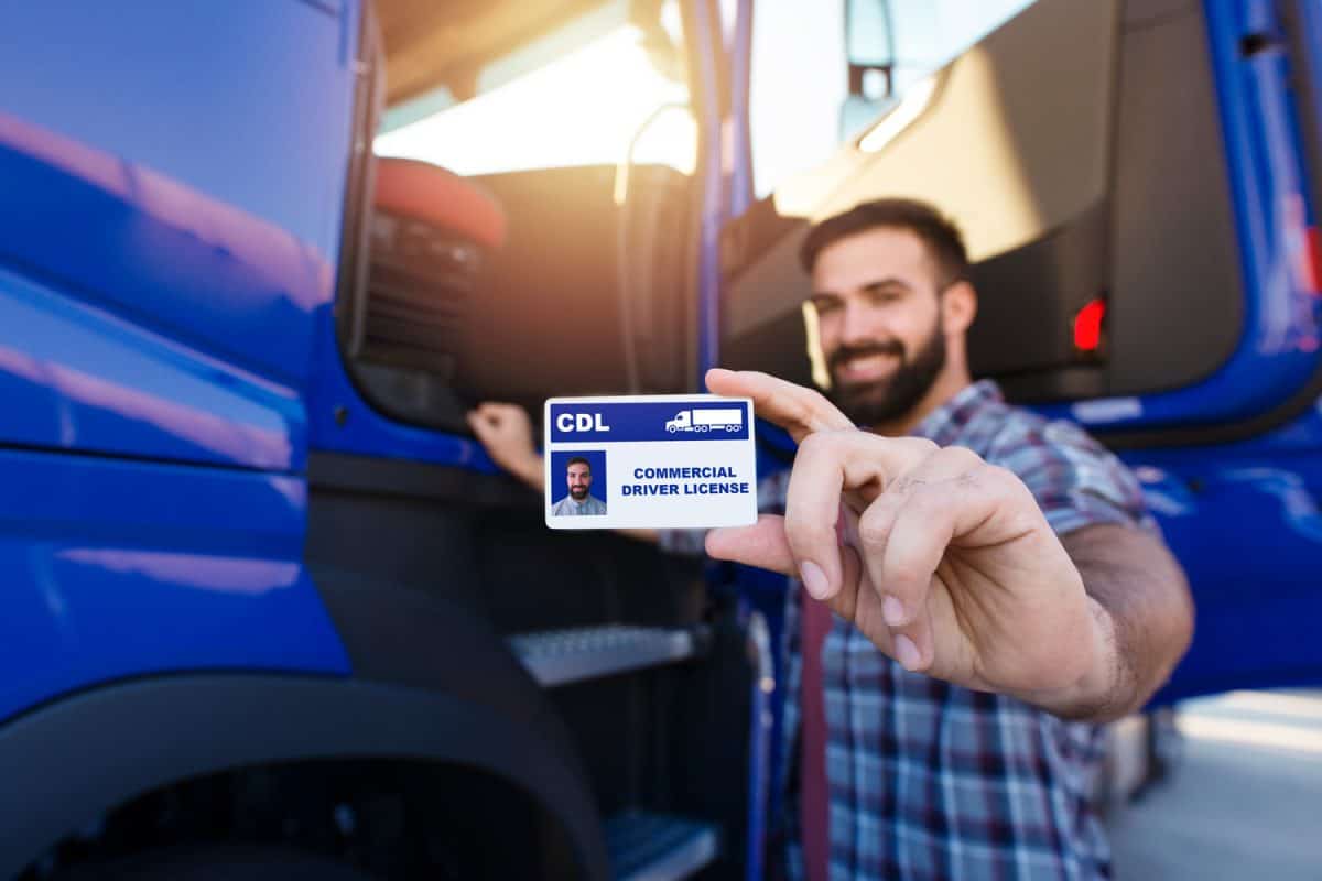 Portrait of middle aged bearded truck driver standing by the truck and showing his commercial driver license. Focus on CDL license. Truck driving school and job openings.
