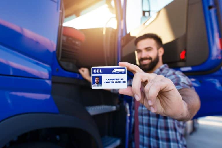 Portrait of middle aged bearded truck driver standing by the truck and showing his commercial driver license. Focus on CDL license. Truck driving school and job openings, Do You Need A CDL To Drive A Box Truck [By State]